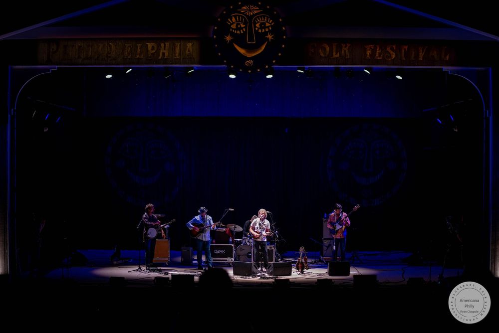 Four musicians performing on a dimly lit stage at the Philadelphia Folk Festival.