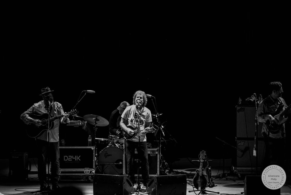 Black and white photo of a band performing on stage with guitars and drums under dramatic lighting.