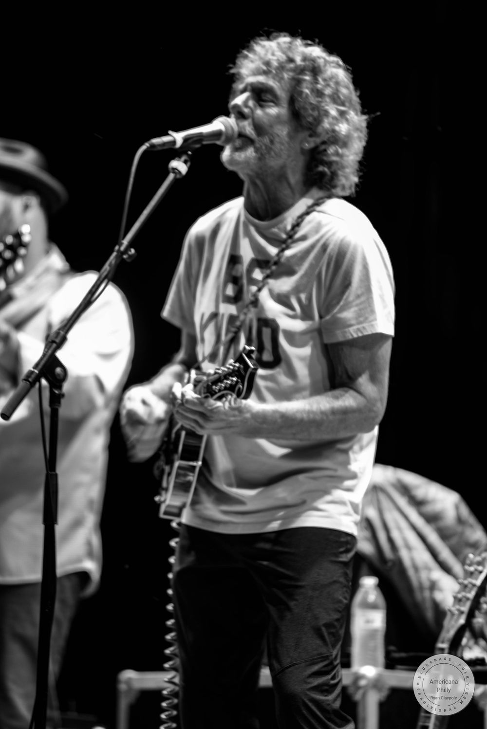 Black and white photo of a curly-haired male guitarist singing into a microphone on stage during a live performance.
