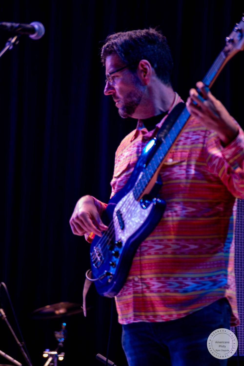 Musician playing electric bass guitar on stage, wearing a colorful striped shirt under concert lighting.