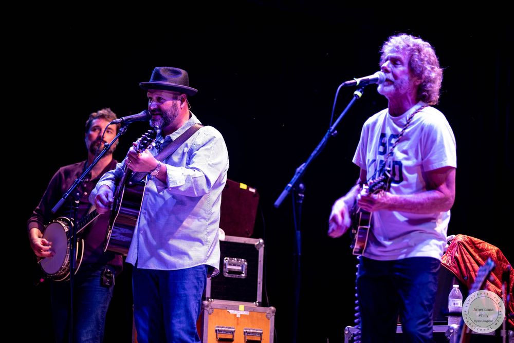 Three musicians perform on stage, playing banjo and guitars at a live concert under warm stage lighting.