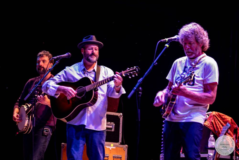 Three musicians performing on stage, two playing acoustic guitars and one electric guitar, under warm stage lighting.