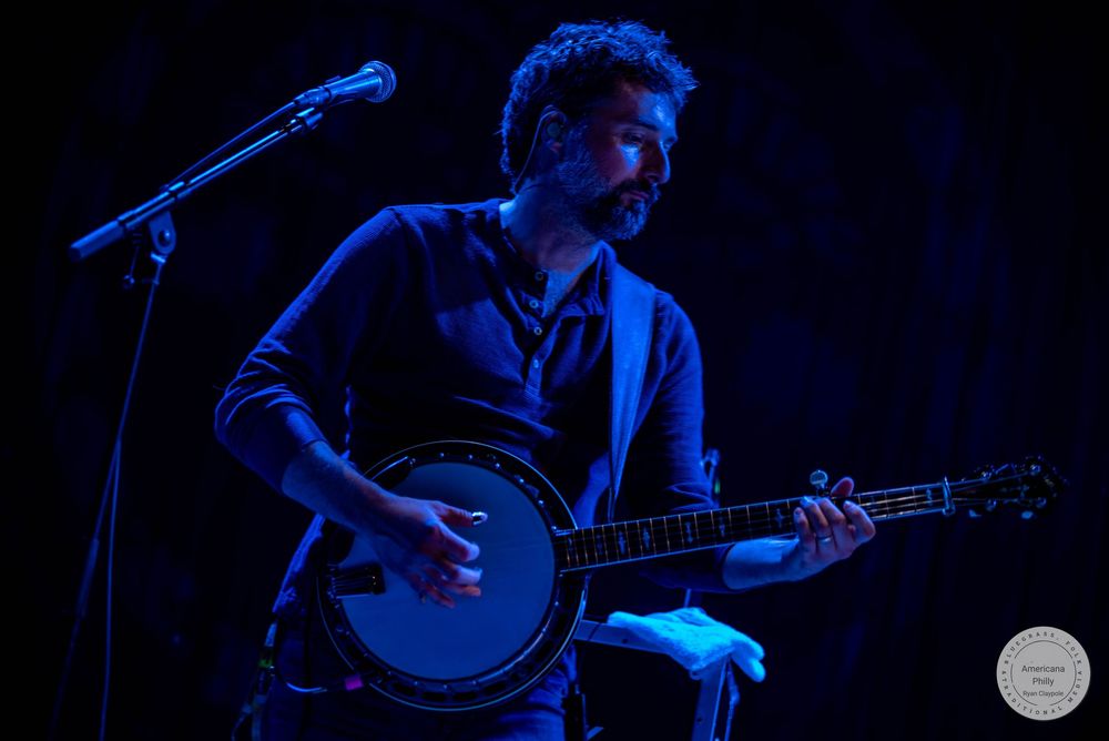 Man playing banjo on stage under blue lighting at a live music concert performance.