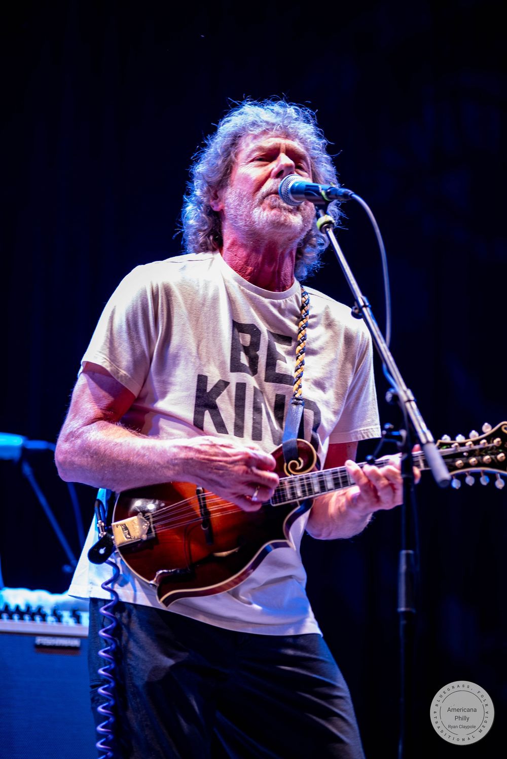 Older male musician playing mandolin on stage, wearing a white Be Kind t-shirt, singing into a microphone at a live concert.
