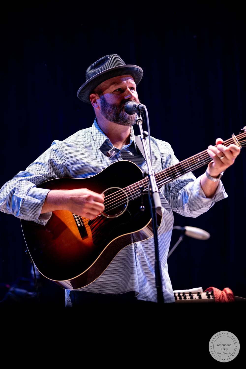Bearded male musician wearing a gray hat performs on stage, singing into a microphone while playing an acoustic guitar.