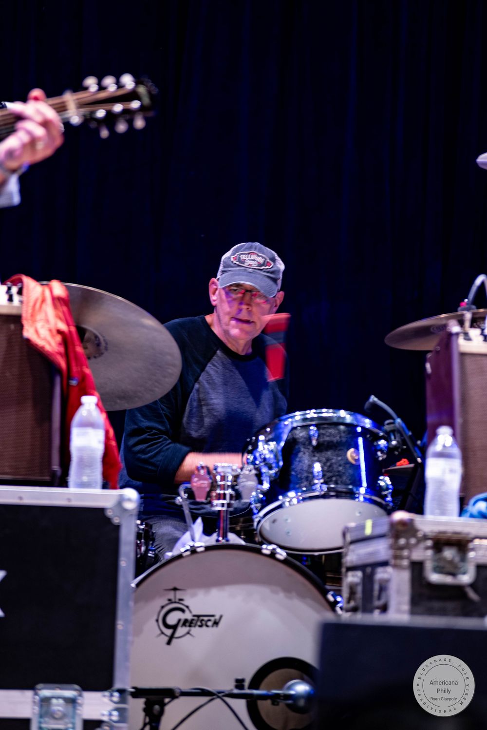 Drummer performing on a Gretsch drum kit on stage, with cymbals and a guitarist visible in the background.