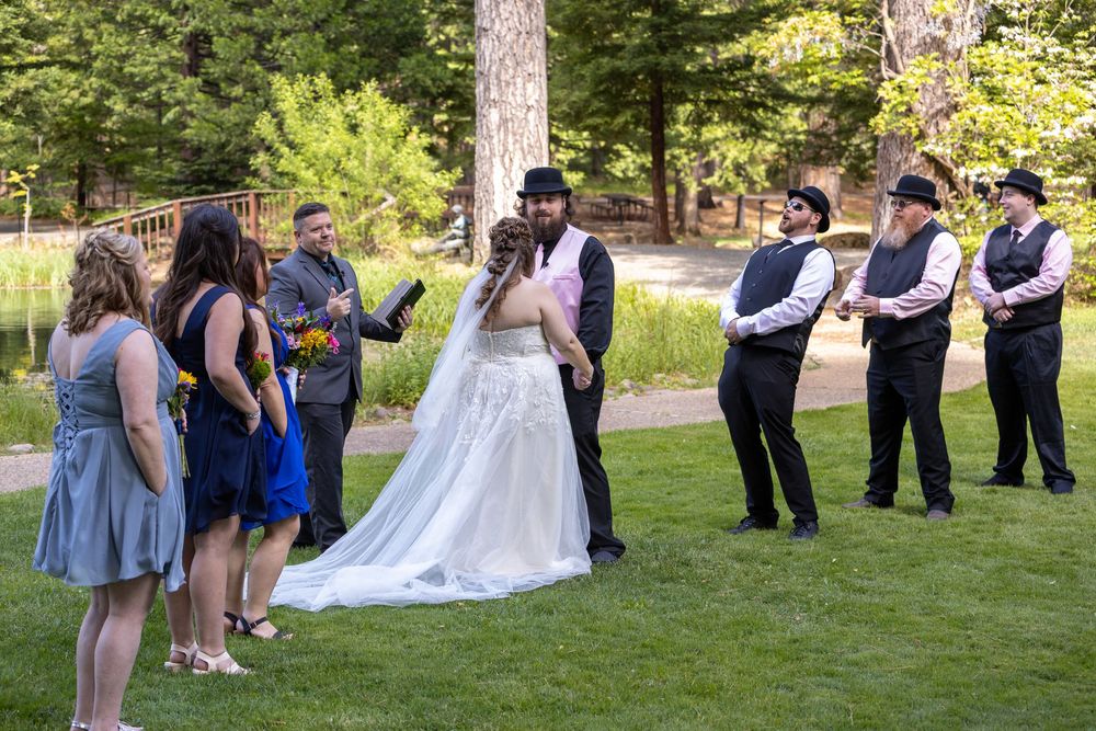 Wedding ceremony outdoors with bride, groom, and wedding party on green lawn among trees.