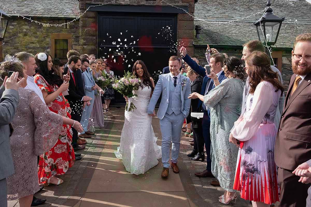 Bride and groom walk through confetti shower, guests lining path outside the Hayloft  at New House Farm wedding venue Lorton