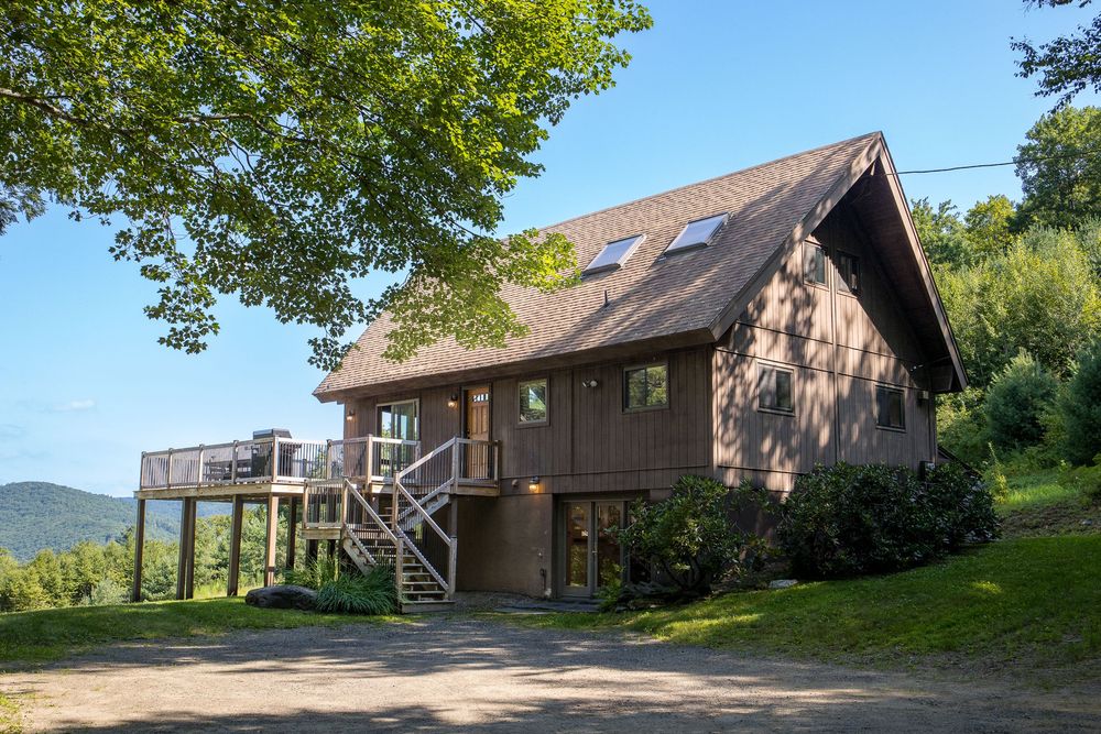 Brown A-frame chalet exterior with deck, skylights, and mountain backdrop on sunny day.