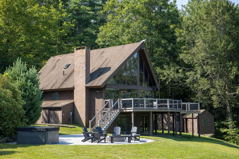 A-frame chalet home with large windows, wood deck, and outdoor seating area surrounded by lush forest.