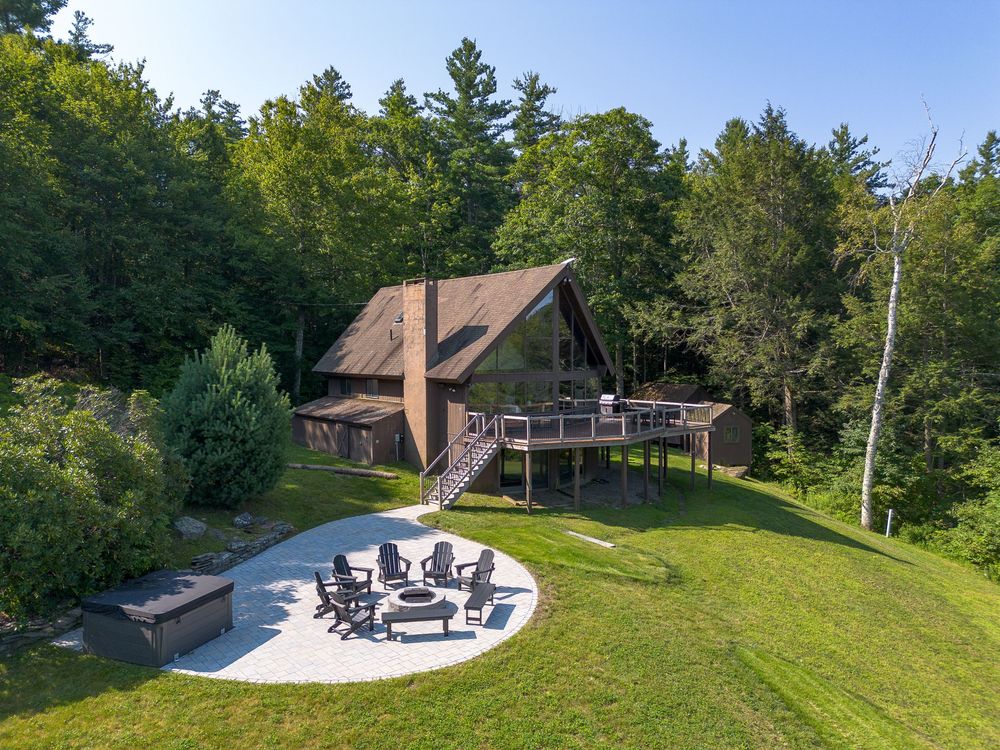 Aerial view of A-frame mountain home with fire pit patio, Adirondack chairs, and verdant lawn.