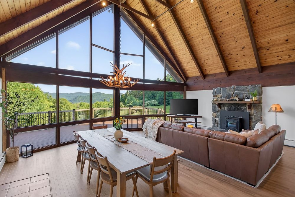 Cathedral ceiling great room with wood beams, floor-to-ceiling windows, stone fireplace, and mountain views.