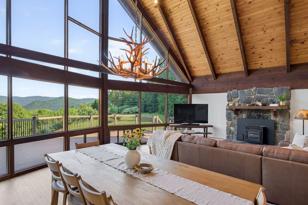 Bright dining area with antler chandelier, large windows, mountain views, and stone fireplace.