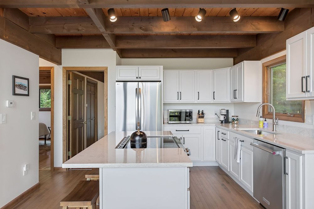 Modern white kitchen with island, stainless steel appliances, and exposed wood beam ceiling.