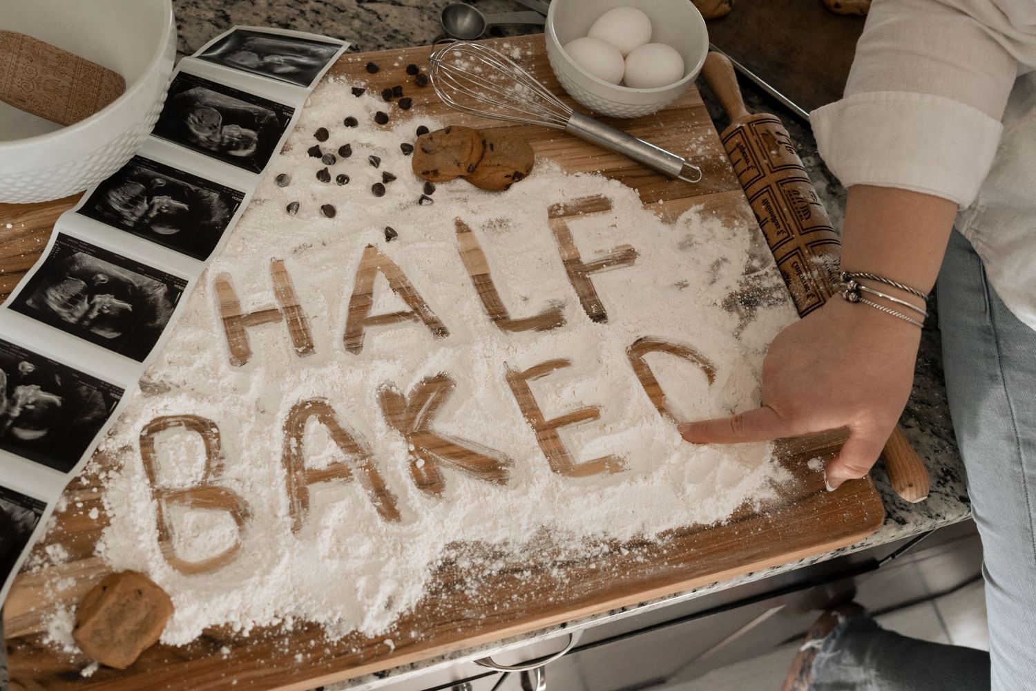 Flour spelling 'Half Baked' on wooden board with eggs, rolling pin, and ultrasound photos for pregnancy announcement.
