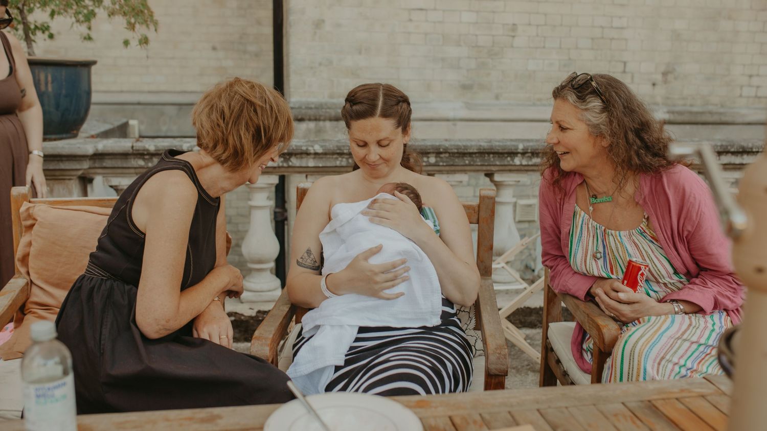 Three women sitting outdoors, one cradling a newborn baby wrapped in white, sharing a warm moment together.