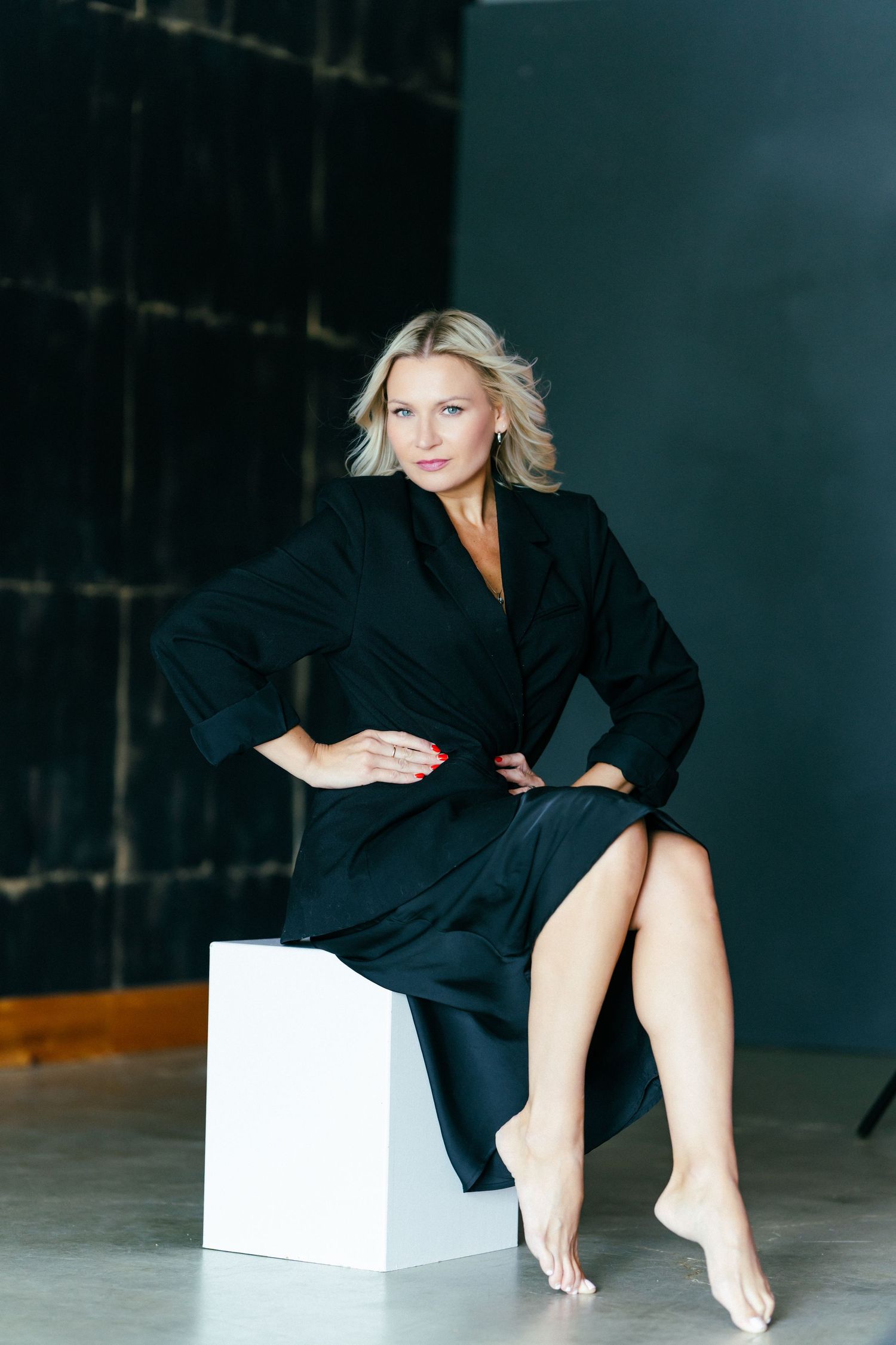 Blonde woman in black blazer dress sits confidently with hands on hips against dark background.
