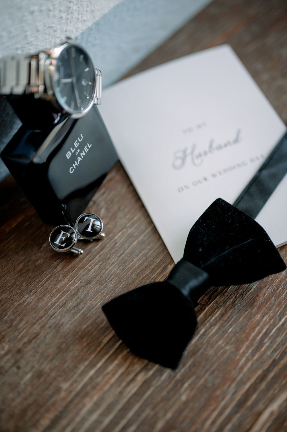 Groom wedding flat lay with black bow tie, Chanel cologne, silver watch, monogram cufflinks, and husband card on wood.