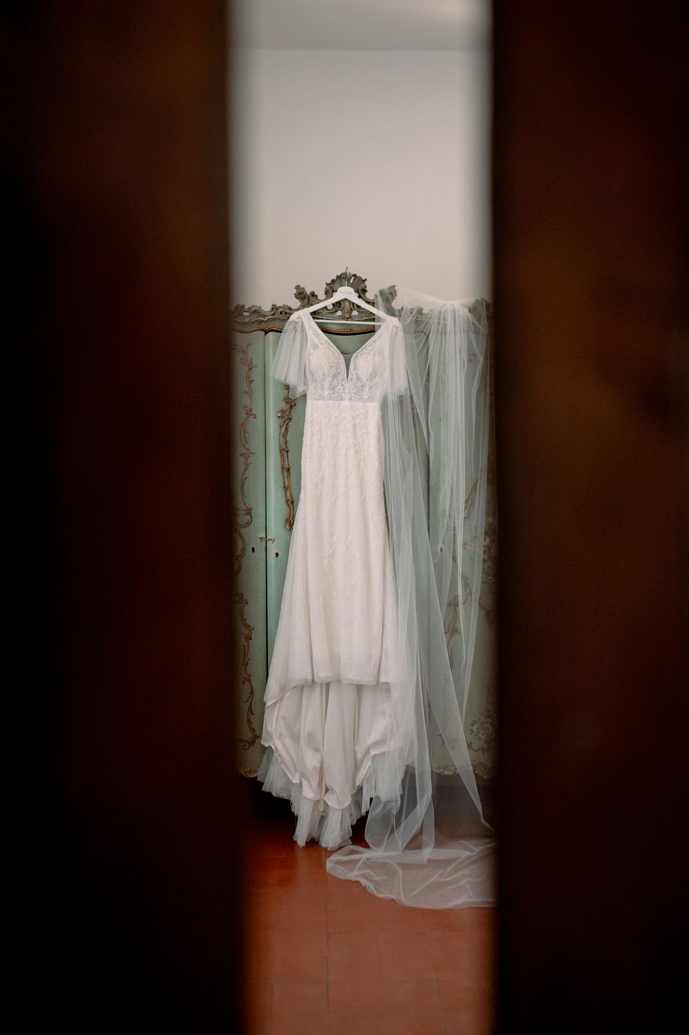 White wedding dress with short sleeves and veil hanging on ornate hanger, viewed through doorway in elegant room.
