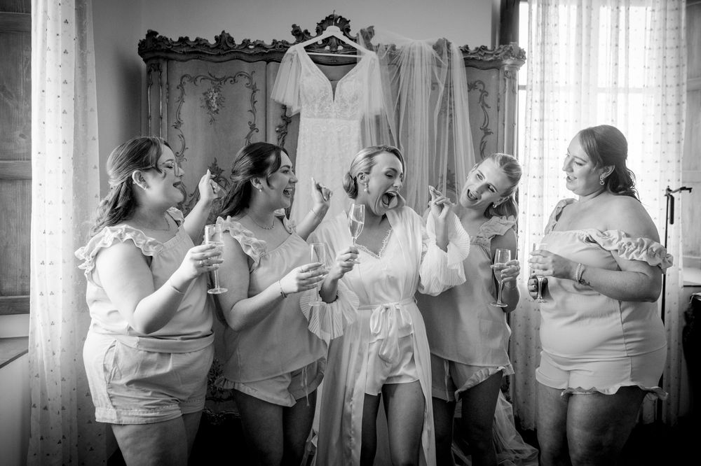 Bridesmaids in white robes celebrate joyfully with the bride, wedding dress hanging in background, black and white photo.