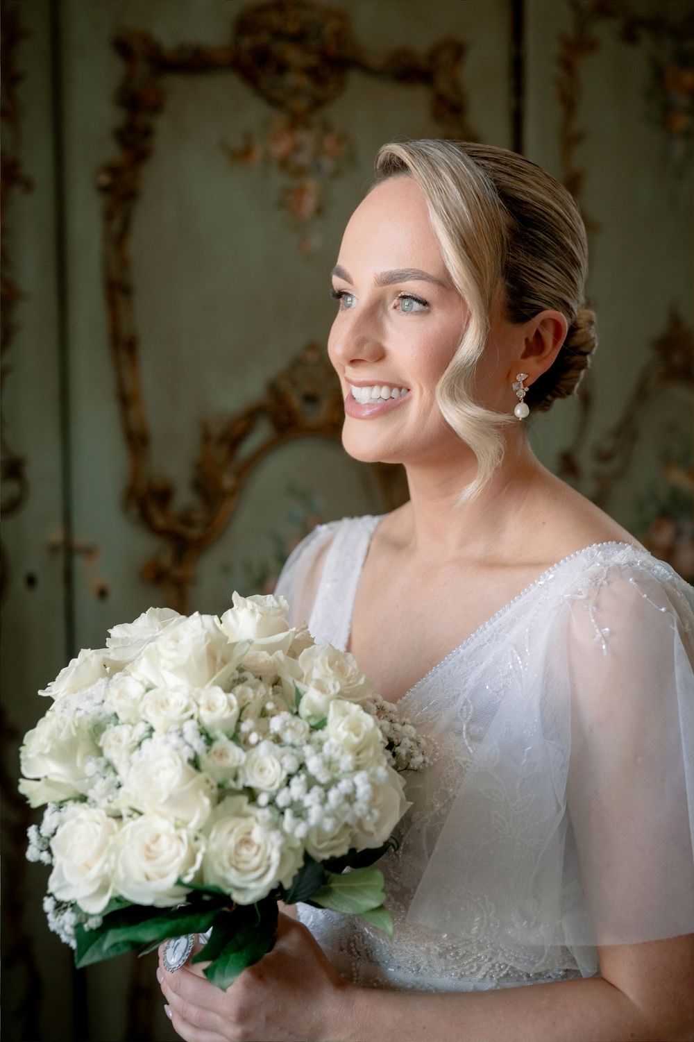 Smiling bride in white lace dress holding elegant bouquet of white roses, with soft curled updo and pearl earrings.
