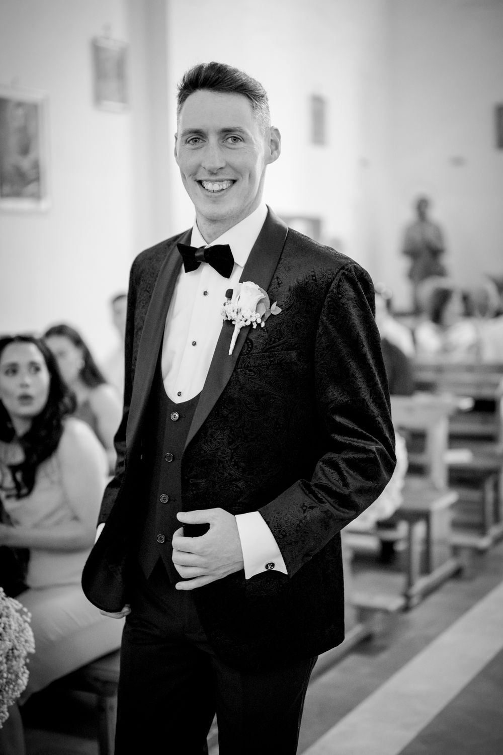 Smiling groom in black tuxedo with boutonniere stands in church aisle on wedding day, black and white photo.