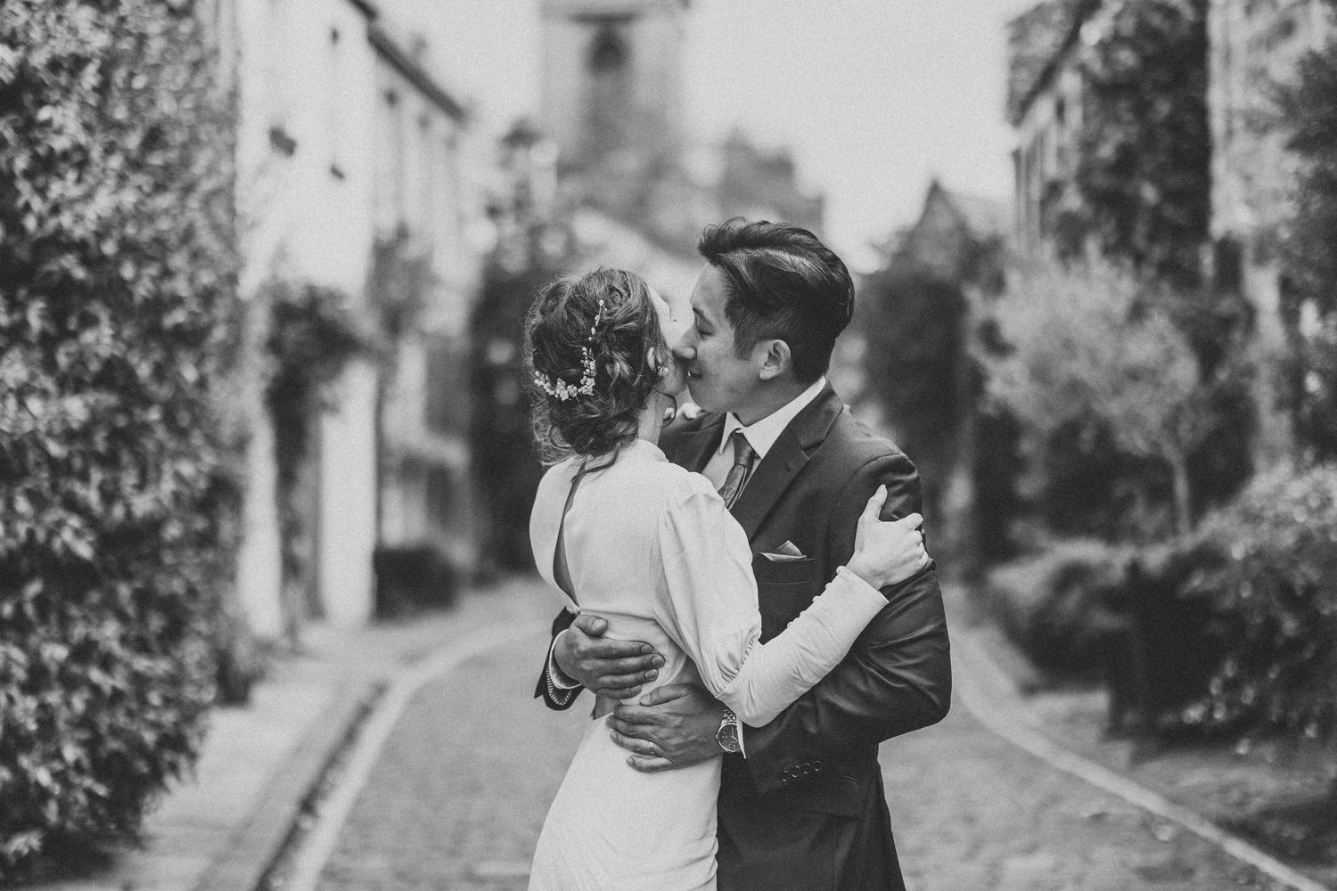 A black and white photo of newlyweds sharing an intimate moment on a vintage street.