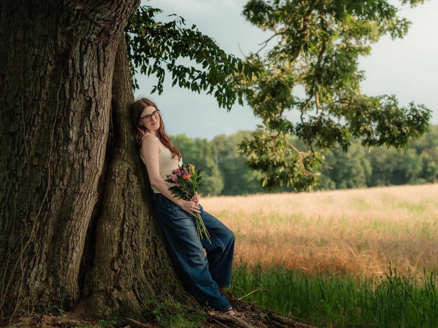 Young woman in jeans leaning against a large tree in a scenic field, holding wildflowers on a sunny day.