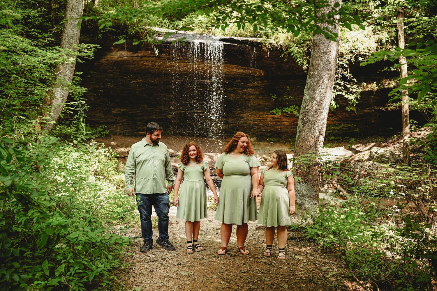 Family of four in matching sage green outfits posing near a waterfall surrounded by lush green forest trees.