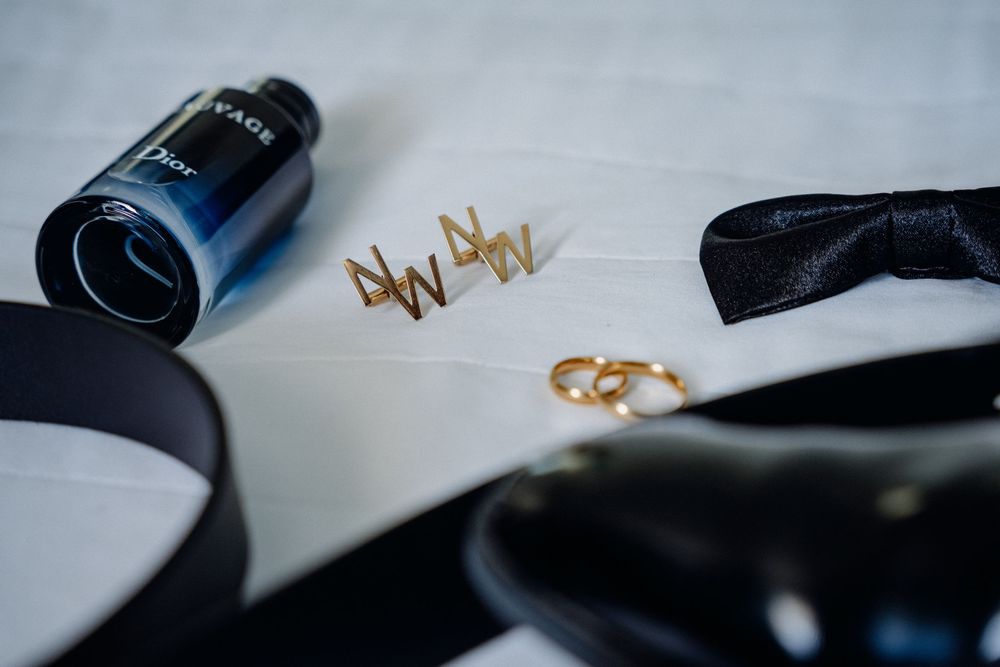 Dior cologne, gold hair pins, wedding rings, black bow tie, and dress shoes arranged on white fabric.