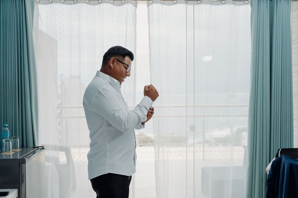 Man in white blazer adjusting cufflinks by sheer curtained window with city view.