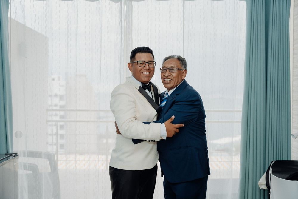 Two men in formal suits smiling and embracing at a wedding celebration near sheer curtained windows.