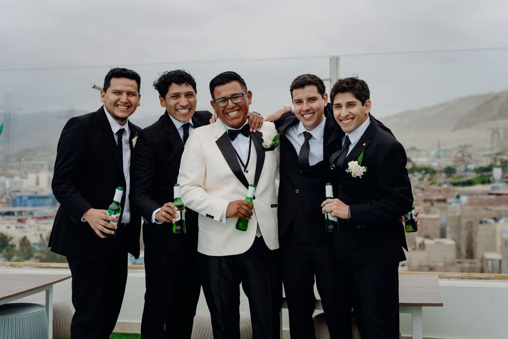 Groom in white tuxedo jacket with groomsmen in black suits holding drinks on rooftop.