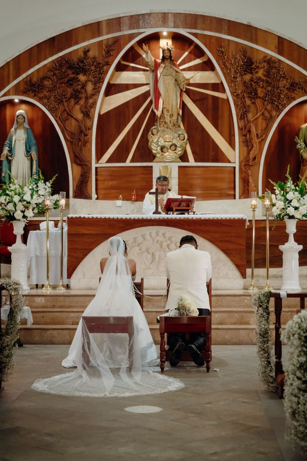 Bride and groom kneel at church altar during Catholic wedding ceremony with floral decorations and religious statue.