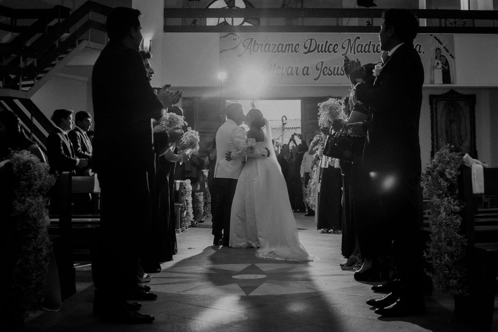 Black and white wedding photo of couple kissing at church altar, guests lining the aisle, sunlight streaming in.
