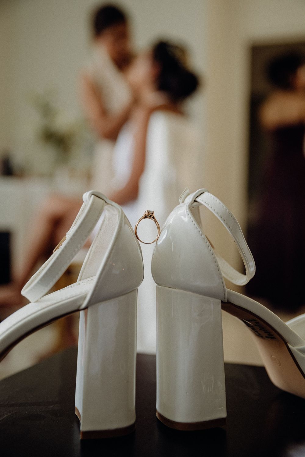 White bridal heels with engagement ring between them, couple embracing in soft focus background.