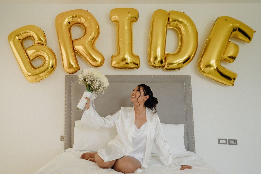 Bride in white robe sits on bed holding bouquet under gold BRIDE balloon letters in bright hotel room.