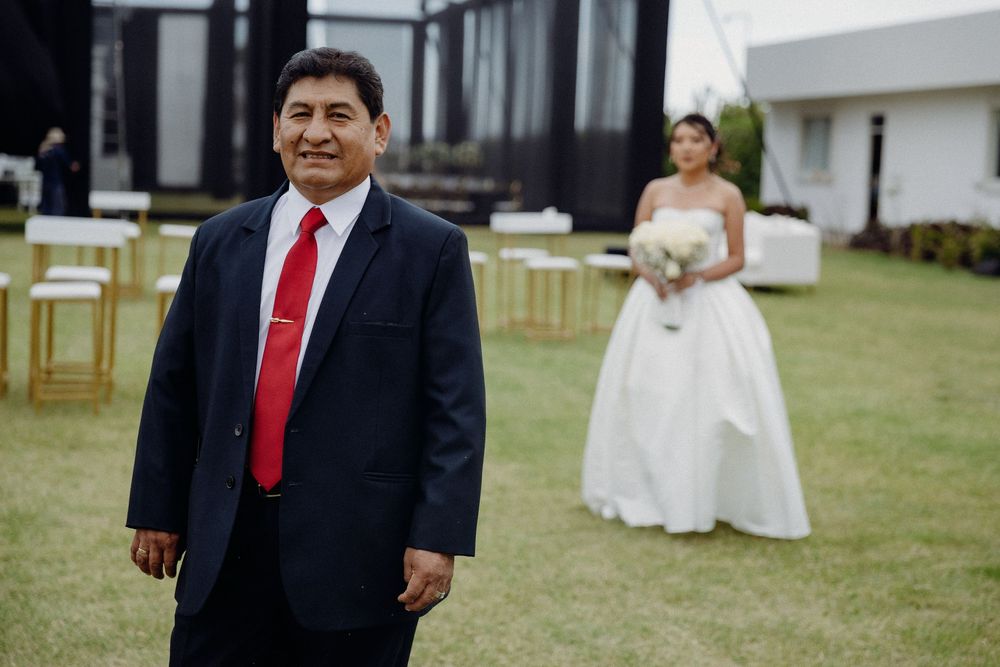 Man in navy suit with red tie stands before bride in white gown holding bouquet at outdoor wedding ceremony.