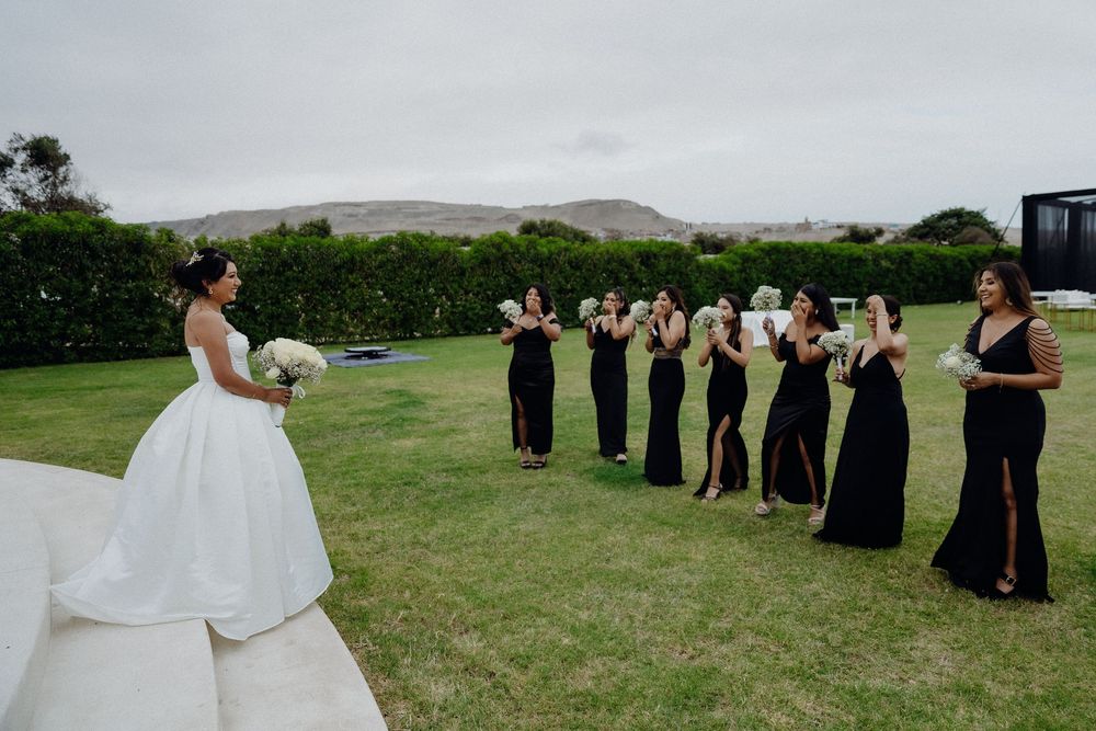 Bride in white gown faces bridesmaids in black dresses holding bouquets on green lawn with hills backdrop.