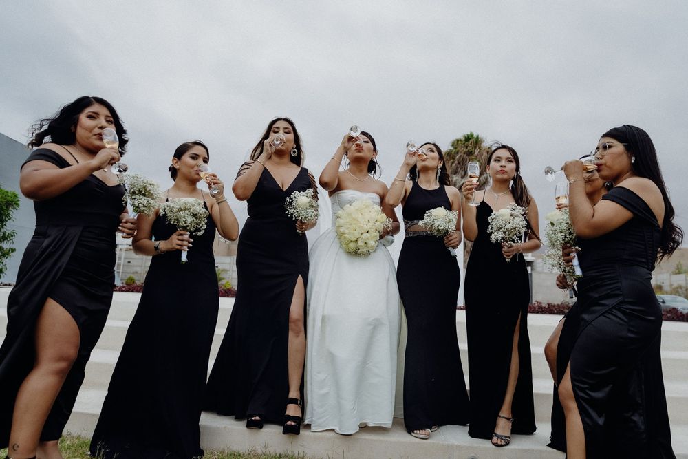Bride in white gown with bridesmaids in black dresses toasting champagne outdoors at a wedding celebration.