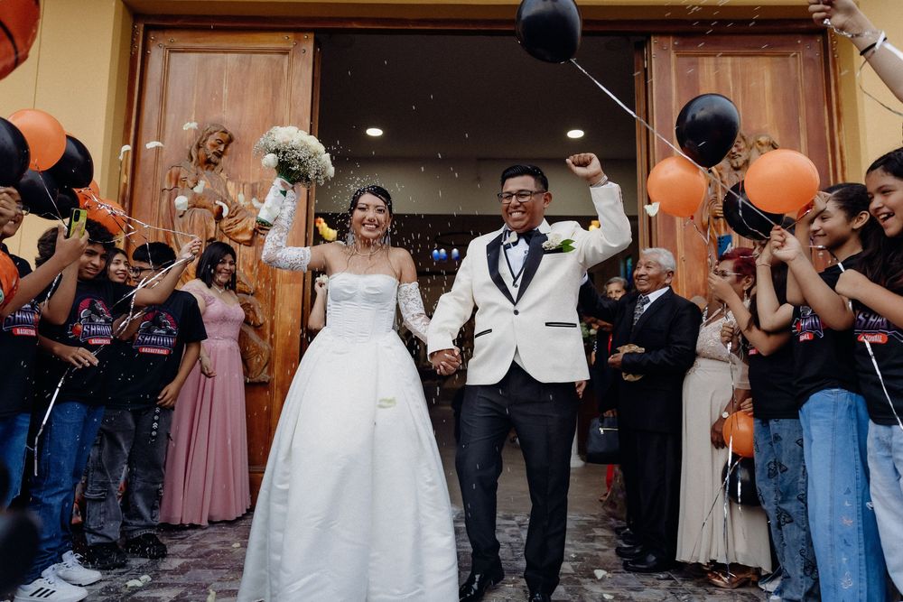 Joyful newlywed couple exits venue in wedding attire, greeted by cheering guests with orange and black balloons.