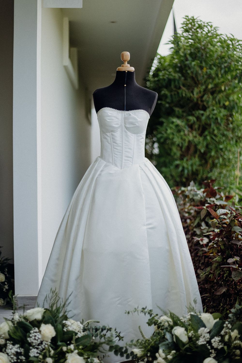 Elegant strapless white ball gown wedding dress displayed on a black mannequin outdoors with green floral arrangements.