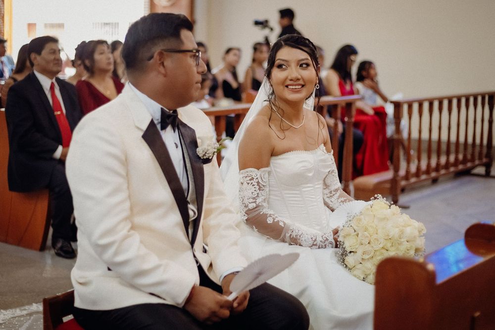 Bride and groom seated at church altar during wedding ceremony, bride wearing strapless lace gown holding white bouquet.