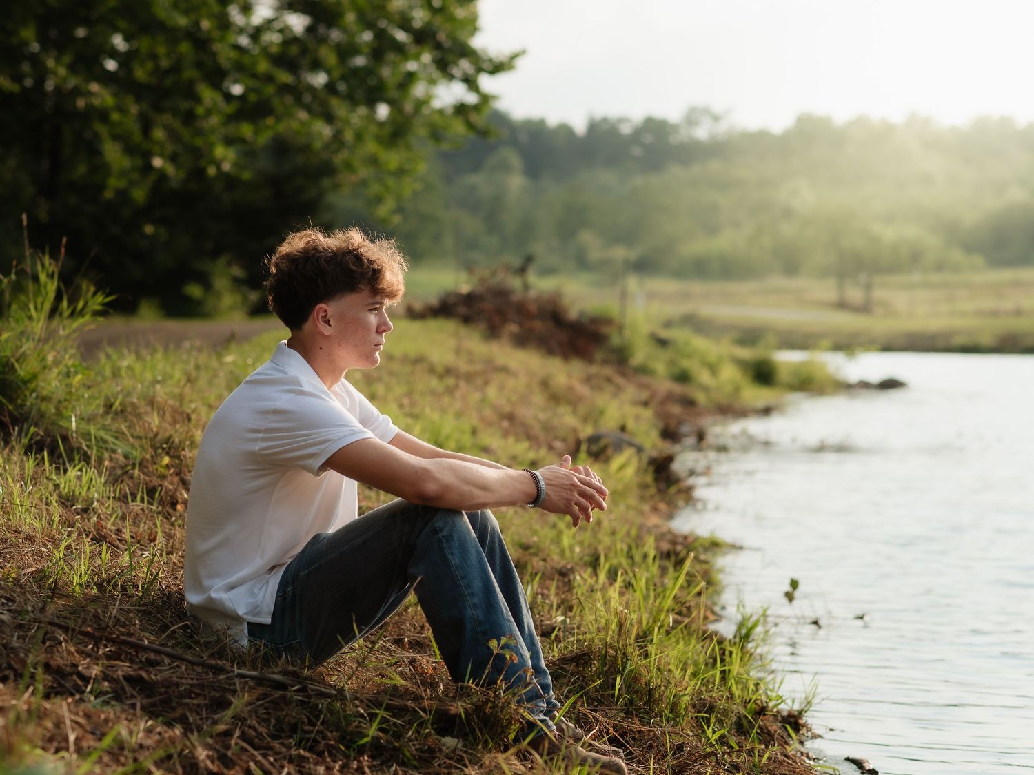 Young man sitting alone by a peaceful lakeside on a sunny day, surrounded by lush green trees and grass.