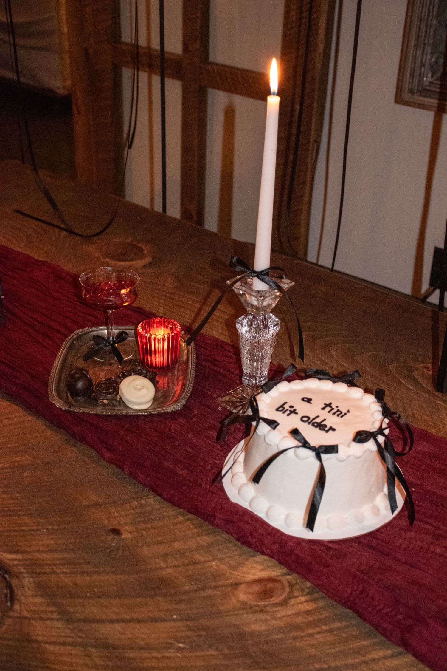 White birthday cake reading a tini bit older beside lit candle and chocolates on red table runner.