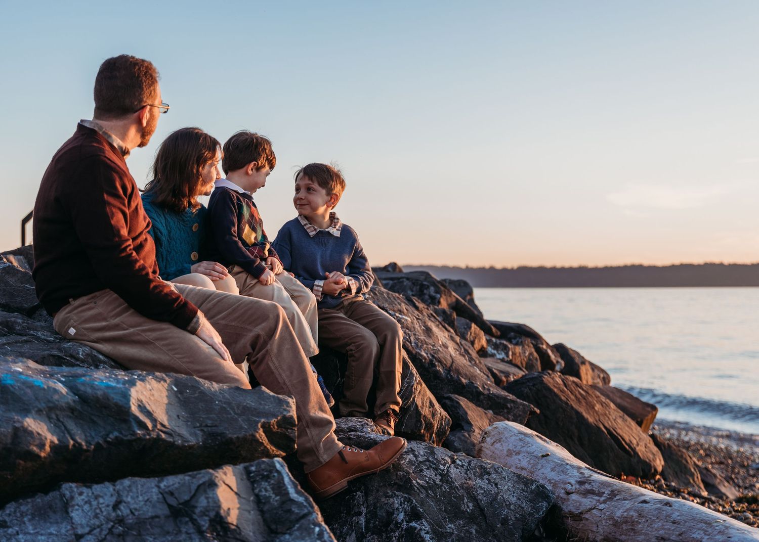 Family of four sits together on large coastal rocks overlooking calm water during a warm golden sunset.