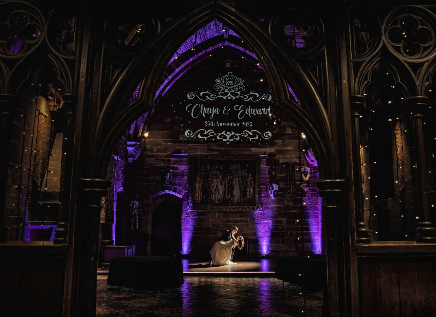 Bride in a white gown dances alone under a gothic stone arch with purple uplighting and a projected monogram at an elegant wedding venue.