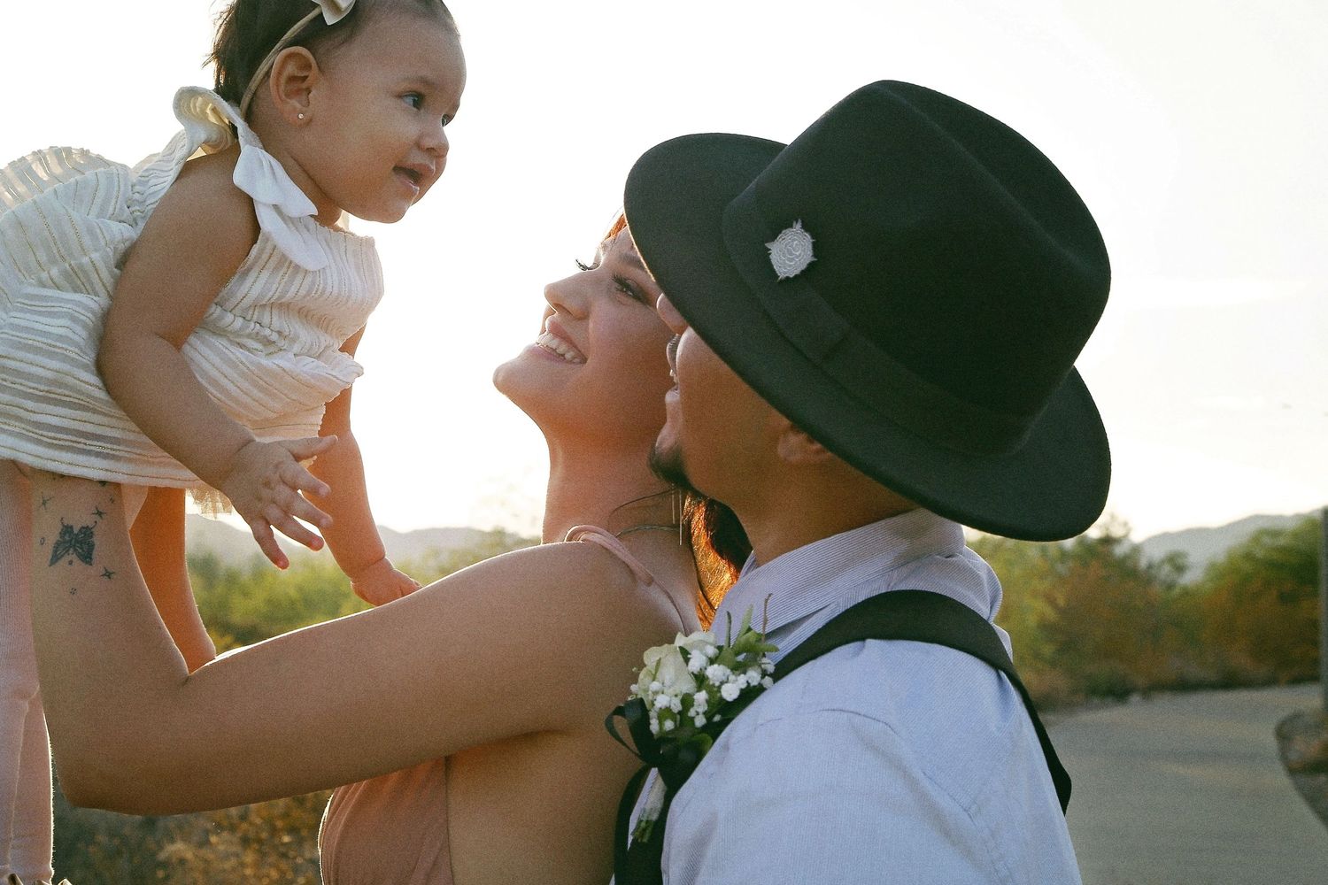 A smiling woman and a man in a black fedora lift a baby in a white dress toward the sky during golden hour.