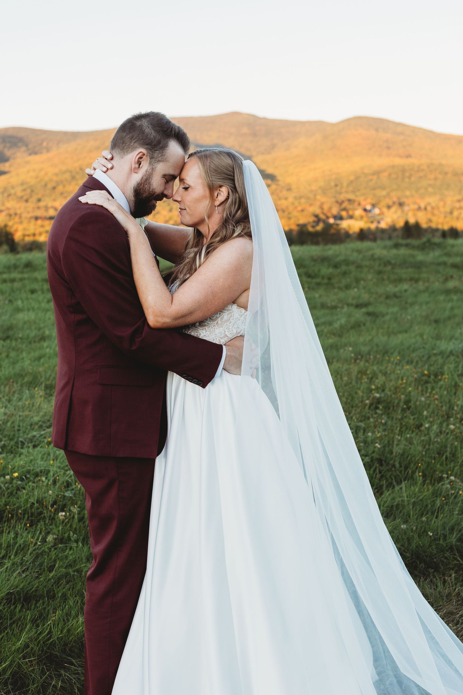 Bride and groom embracing in a field with autumn-colored mountains behind them.