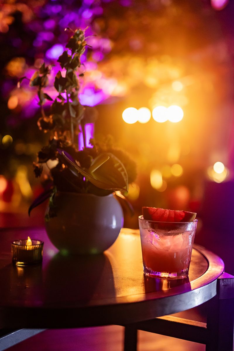 A cocktail with watermelon garnish on a round table beside a vase of flowers and candle in colorful ambient bar lighting.
